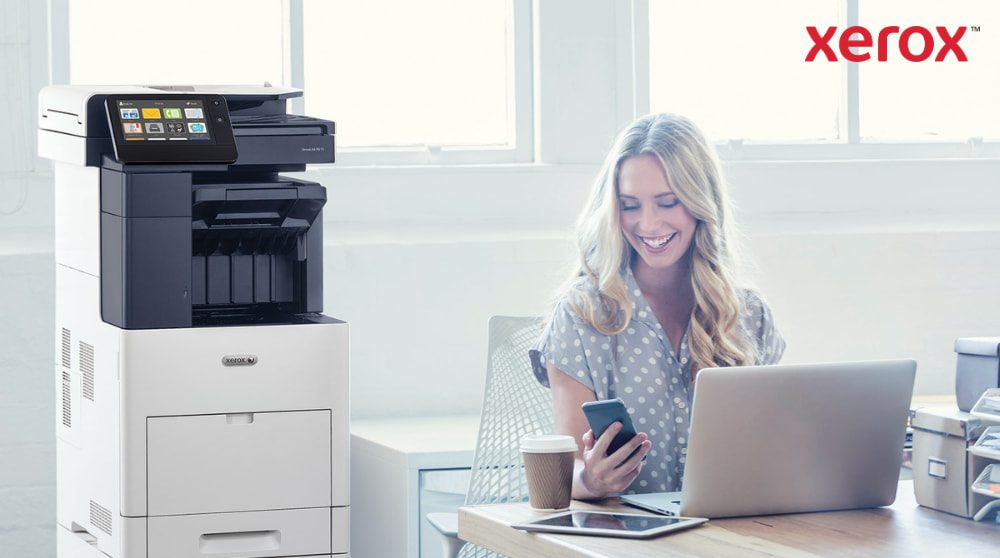 Een persoon kijkt tevreden naar zijn smartphone terwijl hij achter een bureau met een laptop zit. Een Xerox VersaLink B605 multifunctionele printer staat zichtbaar naast het bureau in een helder verlichte kantoorruimte.