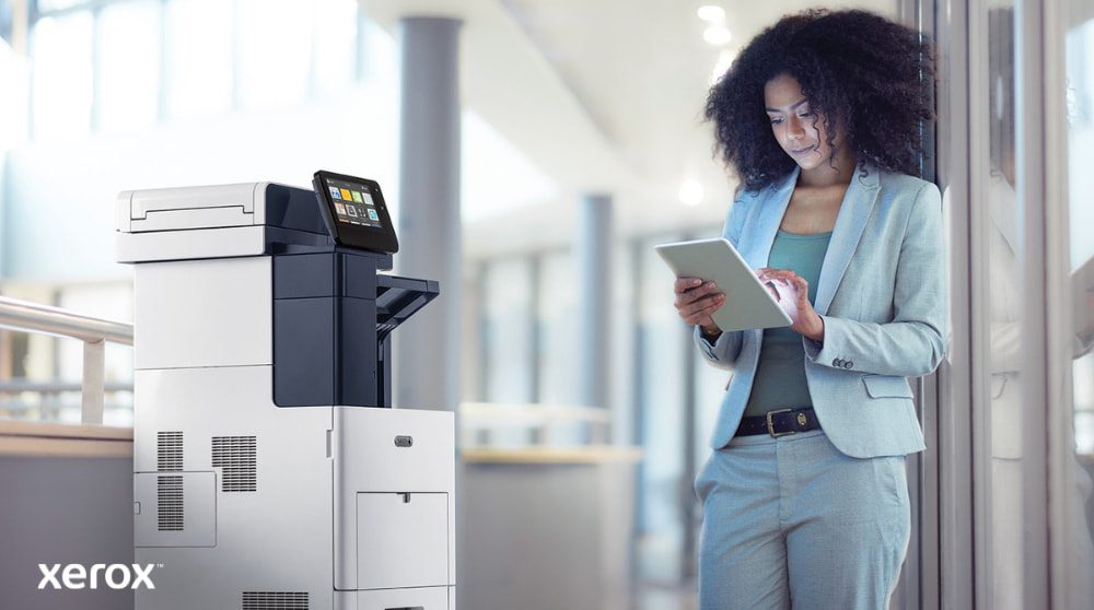 Een persoon staat naast een Xerox VersaLink B605 multifunctionele printer en kijkt naar een tablet. Hij communiceert met deze tablet, wat illustreert hoe ConnectKey mobiele interactie met de printer mogelijk maakt.