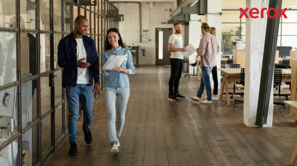 Deux collègues marchent dans un couloir d'un bureau moderne et ouvert, tenant des documents et souriant, avec d'autres collègues visibles en arrière-plan, représentant un lieu de travail dynamique et collaboratif.