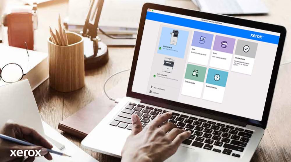 Iemands handen liggen op het toetsenbord van een laptop en op het scherm wordt de interface van Xerox Print and Scan Experience weergegeven, met opties zoals Scannen, Afdrukken, Apparaatstatus, Benodigdheden bestellen en Ondersteuningscentrum, wat wijst op een gebruiksvriendelijke bediening.