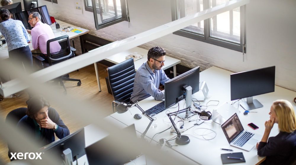 Een bovenaanzicht van een open kantoorruimte met meerdere mensen die achter hun bureau werken en computers gebruiken. De scène straalt een productieve en collaboratieve werkomgeving uit.