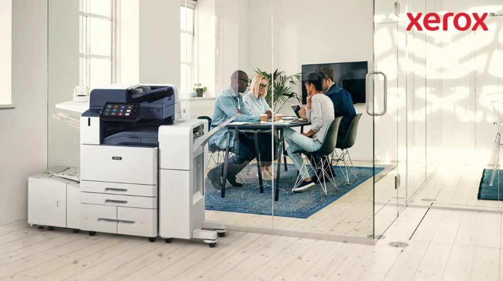 Une imprimante multifonction Xerox AltaLink B8245 moderne trône au premier plan d'un bureau lumineux. À l'arrière-plan, un groupe de personnes discutent autour d'une table.