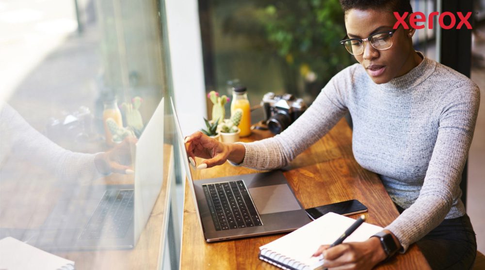 Xerox® Workplace Cloud en Suite: verbeterde productiviteit Een persoon met een bril werkt aan een houten tafel, kijkt naar een laptopscherm en schrijft in een notitieboekje, met een smartphone in de buurt, wat duidt op een productieve werkomgeving